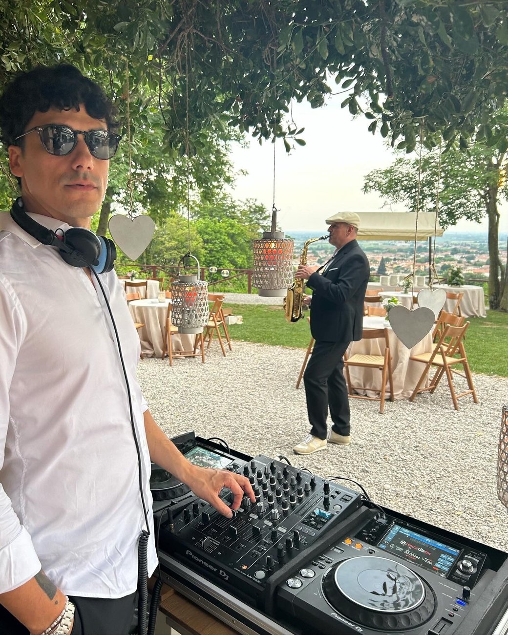 Jam Mattia con cuffie al collo alla console Pioneer durante un evento all'aperto a Treviso, sassofonista sullo sfondo nel giardino