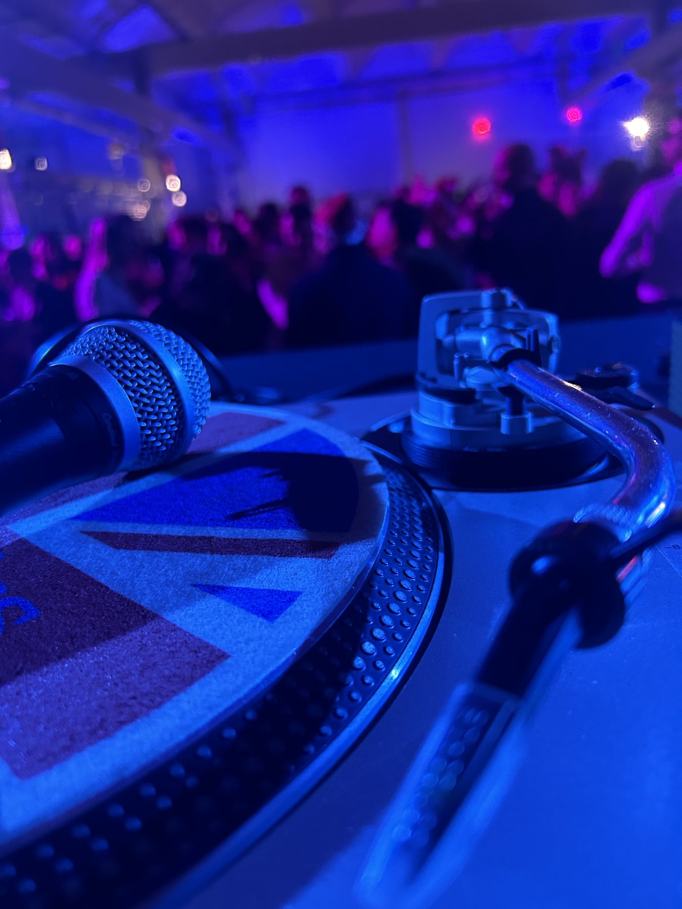 Microfono e giradischi in primo piano con luce blu, pubblico di una festa aziendale sullo sfondo durante il DJ set