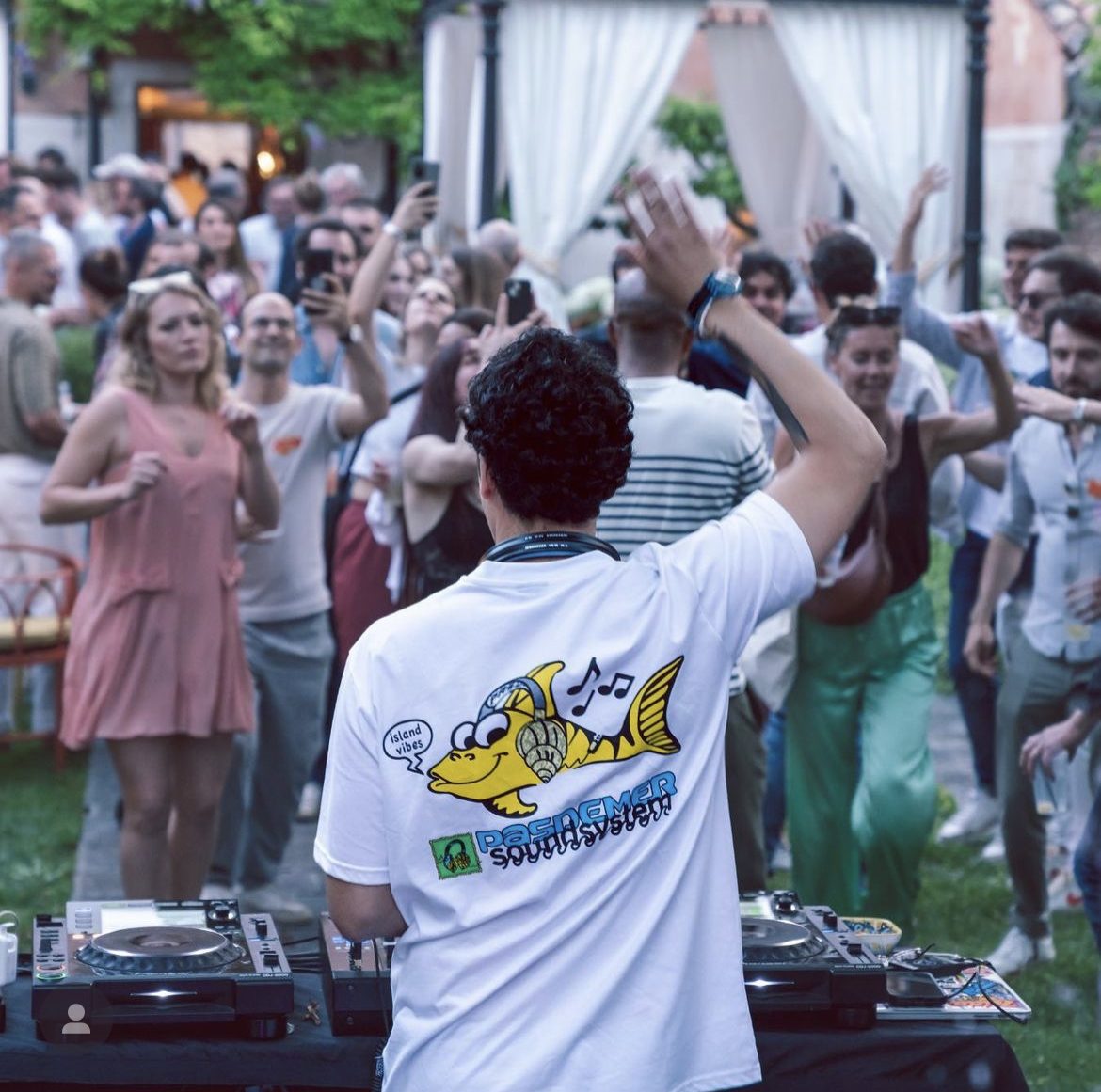 DJ set al Lido di Venezia con ospiti internazionali che ballano a braccia alzate davanti alla console nel giardino: festa di matrimonio con pubblico coinvolto