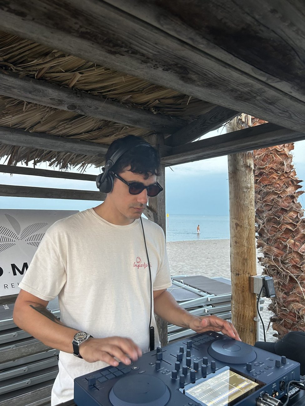 Jam Mattia in DJ set al mare sotto un gazebo di paglia con la spiaggia alle spalle, beach party estivo