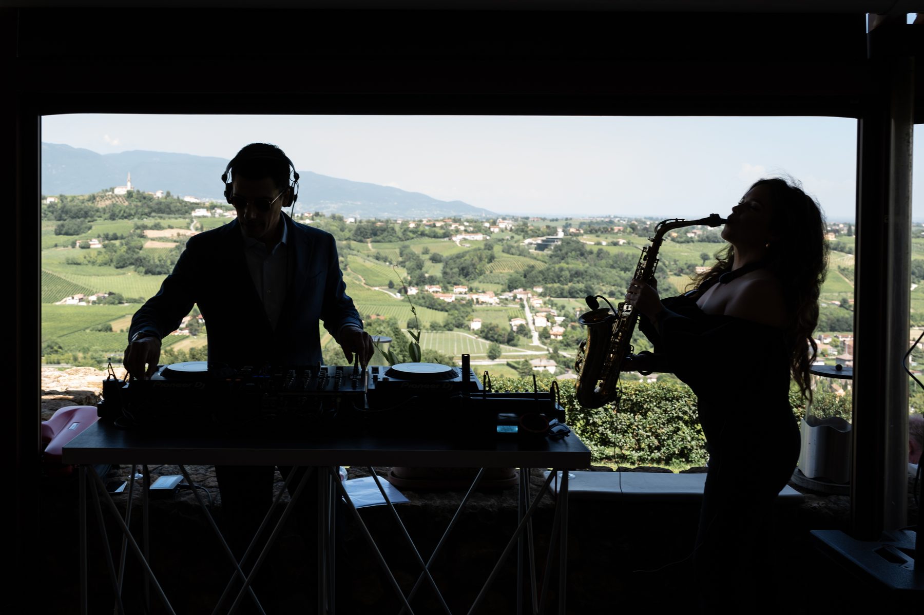 DJ e sassofonista dal vivo in controluce a Ca' del Poggio (Treviso), location immersa nelle colline trevigiane: contrasto tra le sagome scure della postazione e il panorama luminoso sui vigneti