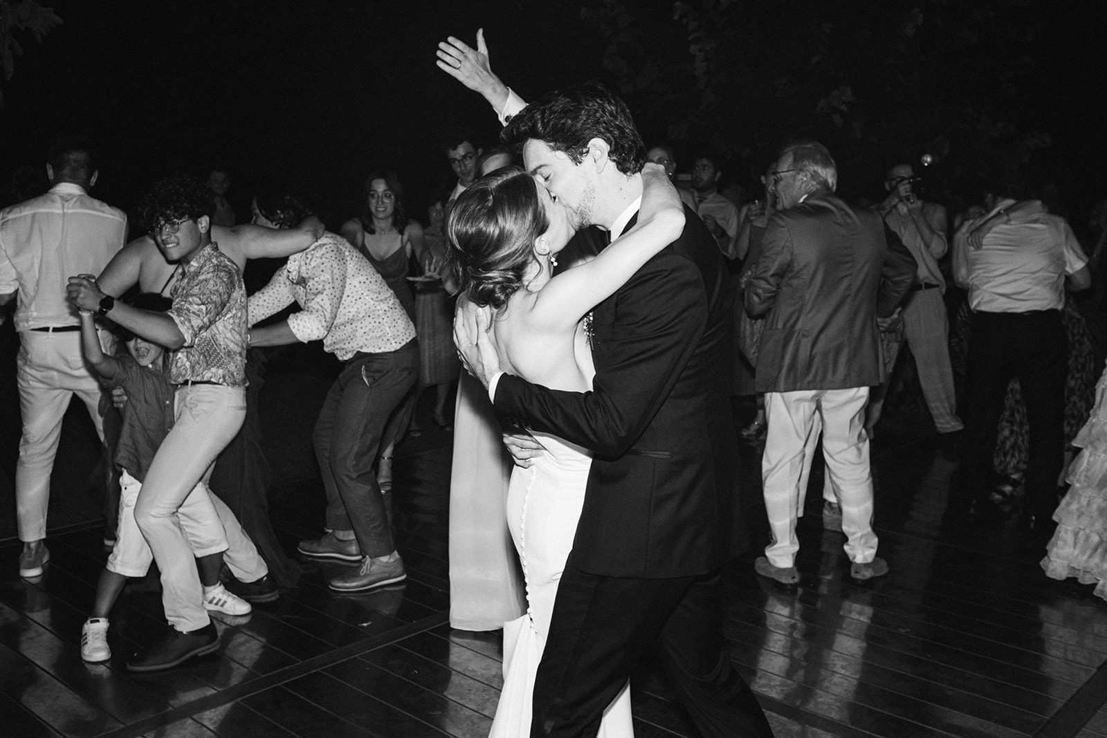 Foto in bianco e nero di un matrimonio: in primo piano il bacio degli sposi, dietro gli invitati che ballano sulla musica del DJ set live di Jam Mattia, non una playlist ma una selezione che si adatta alla serata