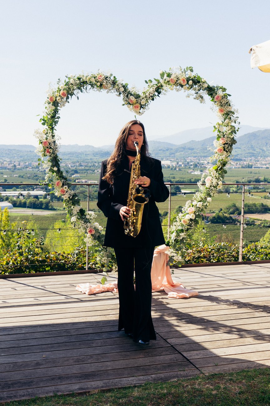 Sassofonista con sax wireless davanti al cuore di fiori sulla terrazza di Castelletto Eventi (Treviso): sax live che accompagna il DJ set di Jam Mattia durante il matrimonio